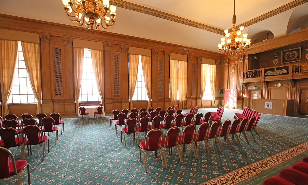 A view of the Banqueting Hall at Leeds Civic Hall set up for a wedding ceremony. The view is from the back of the room, the walls of the room are clad in ornate wooden panelling, large windows run down the walls and two large chandelliers hang from the ceiling
