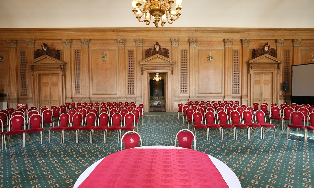 A view of the Banqueting Hall at Leeds Civic Hall set up for a wedding ceremony. The view is from the front of the room, the walls of the room are clad in ornate wooden panelling and a large chandellier hangs from the ceiling aligned in the centre of the photo