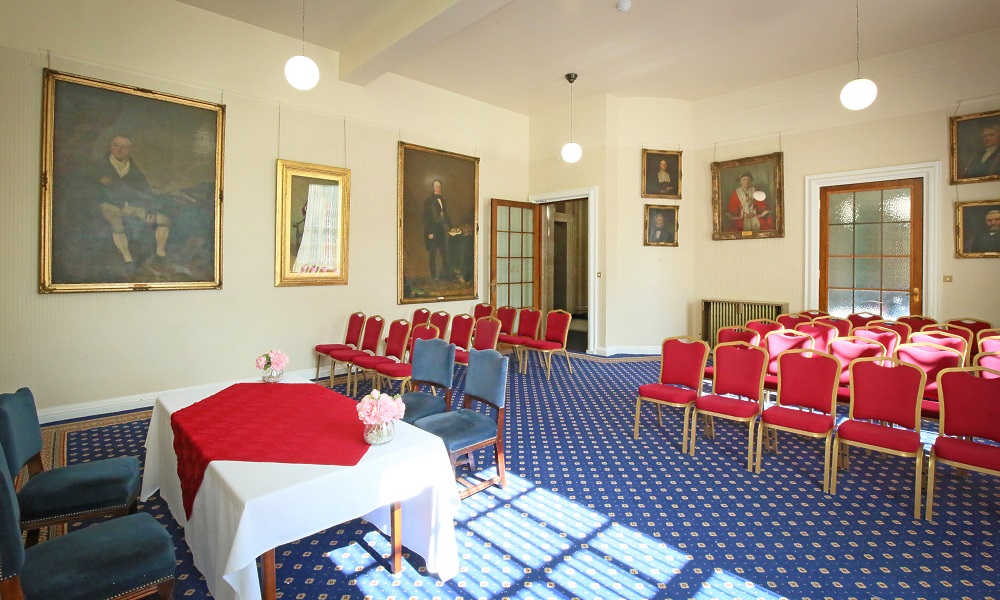 A view of the West Room at Leeds Civic Hall from the front left corner, the walls have historic portraits and there are two large windows with blinds to the right.