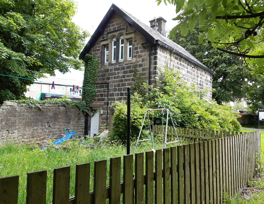 a grey stone detached house with a grass back garden, 4 long thin windows on the first floor, large stone wall bordering a main road and wooden fence bordering Manor Garth Park