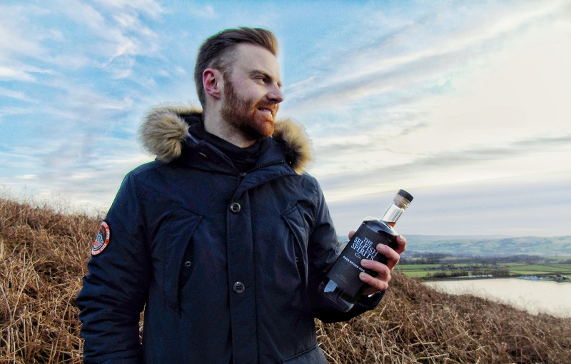 Fred Harman, founder of The Selfish Spirits Co holding a bottle looking out over the moors