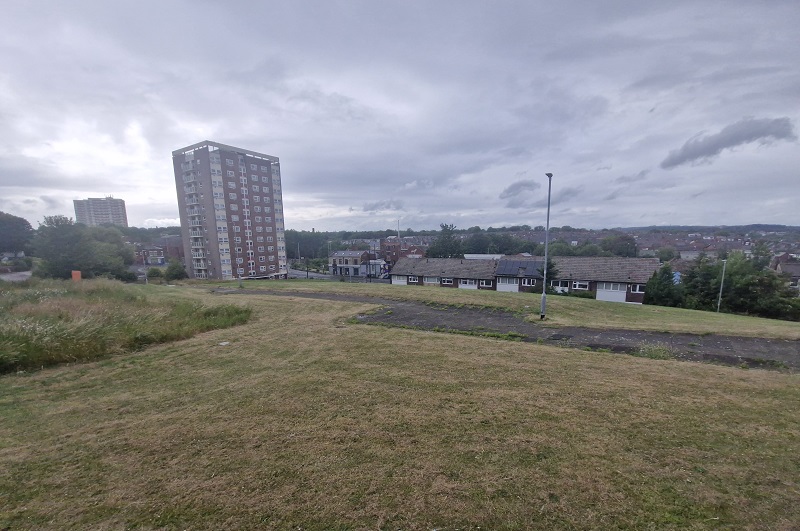 Alternative view of land at Mistress Lane, showing green grassy land raised slightly from the street level, opposite terraced houses and a block of flats.