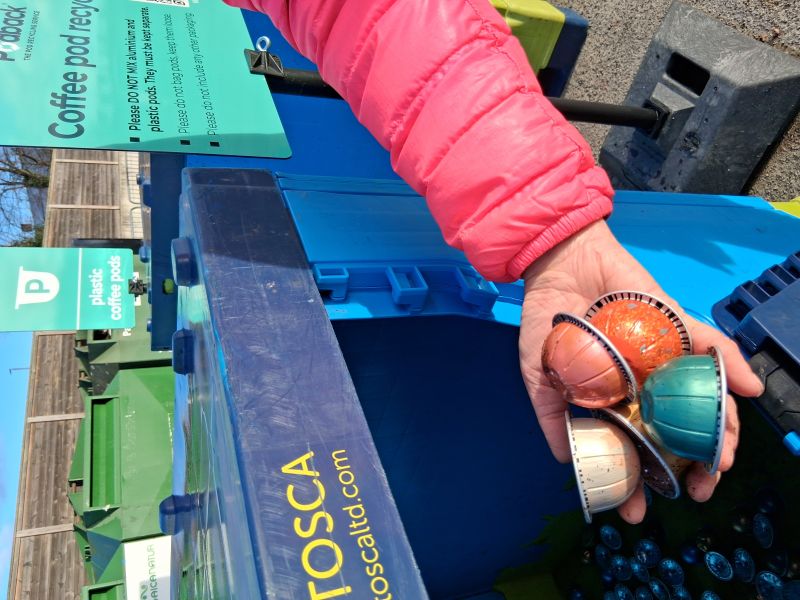 Blue container with hand holding used aluminium coffee pods for recycling 