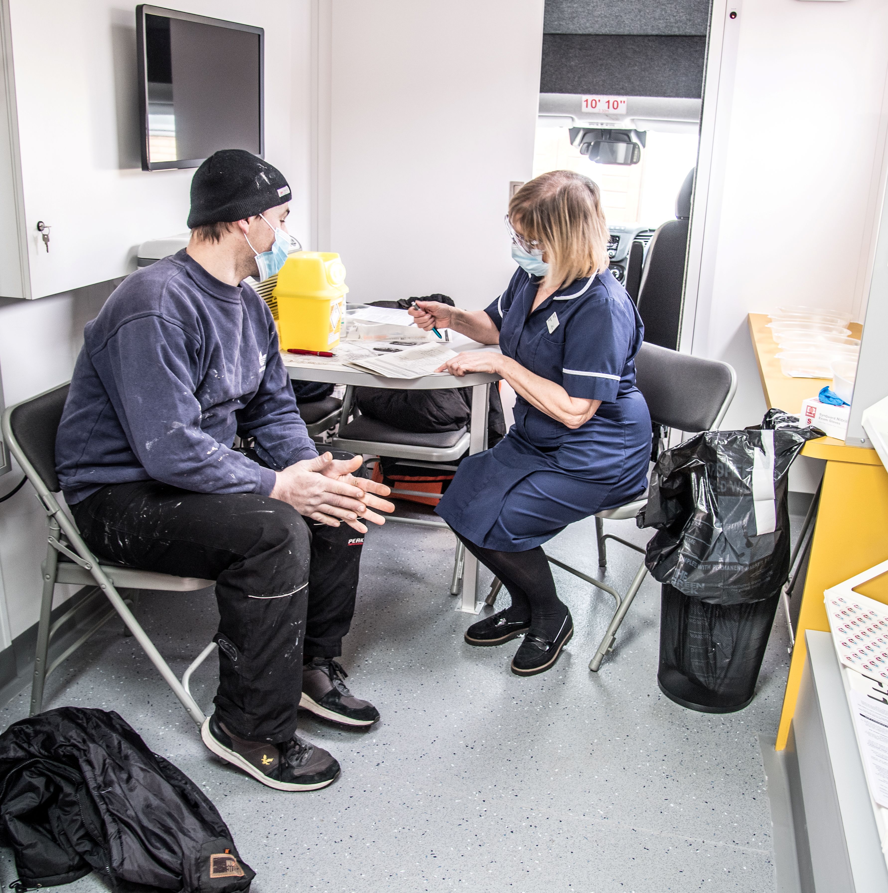 Image shows seated man wearing a facemask in conversation with a seated woman NHS worker wearing a facemask