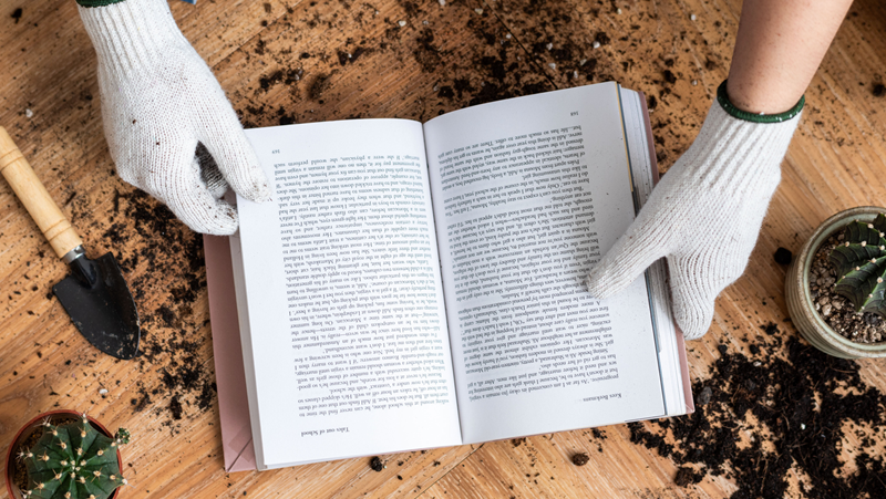 open book surrounded by gardening tools and being opened by hands wearing white gardening gloves