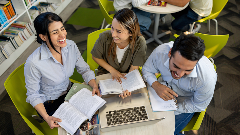 3 students sat in a library with open books and laughing