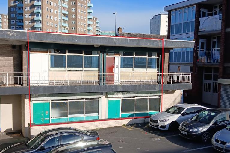 Two storey building with a balcony. six cars are parked in front of the building and a tower block sits behind it surrounded by blue sky