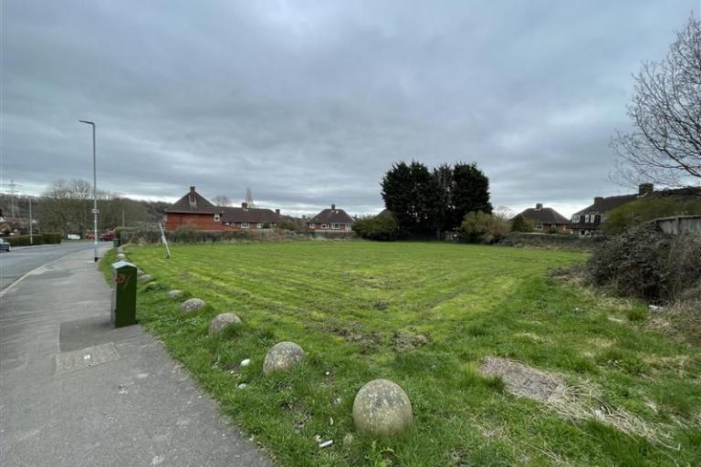 Image shows vacant grassed area next to pavement and bordered by houses